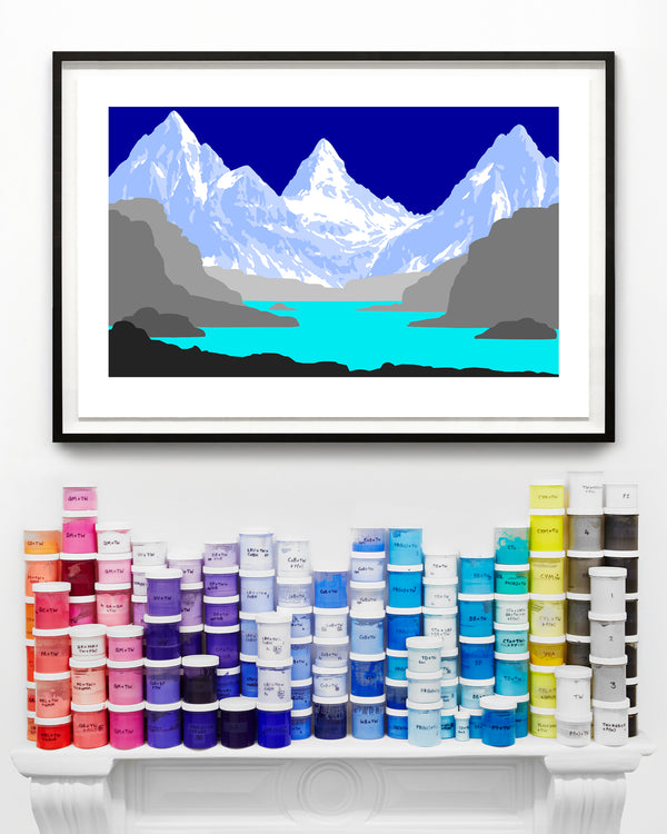 Framed contemporary art print of a mountain landscape with a teal lake and blue sky, displayed above a mantel piece with paint containers.