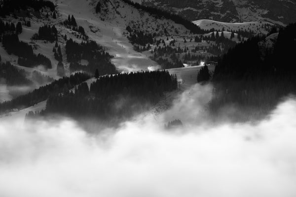 Black and white ski resort photo. A minimalist winter landscape showing snow cannons on groomed mountain slopes above a thick layer of valley fog at dawn.