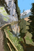 Artistic rendering of a steep mountain path along a cliffside gallery, featuring expressive brushstrokes of green vegetation and grey rock faces under a bright blue alpine sky.