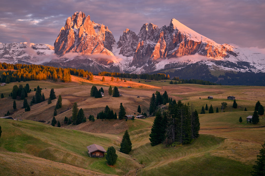 Dolomites art print. A mountain landscape featuring golden rolling meadows and small wooden cabins beneath the jagged peaks of the Italian Alps glowing with sunset light and a fresh dusting of snow.