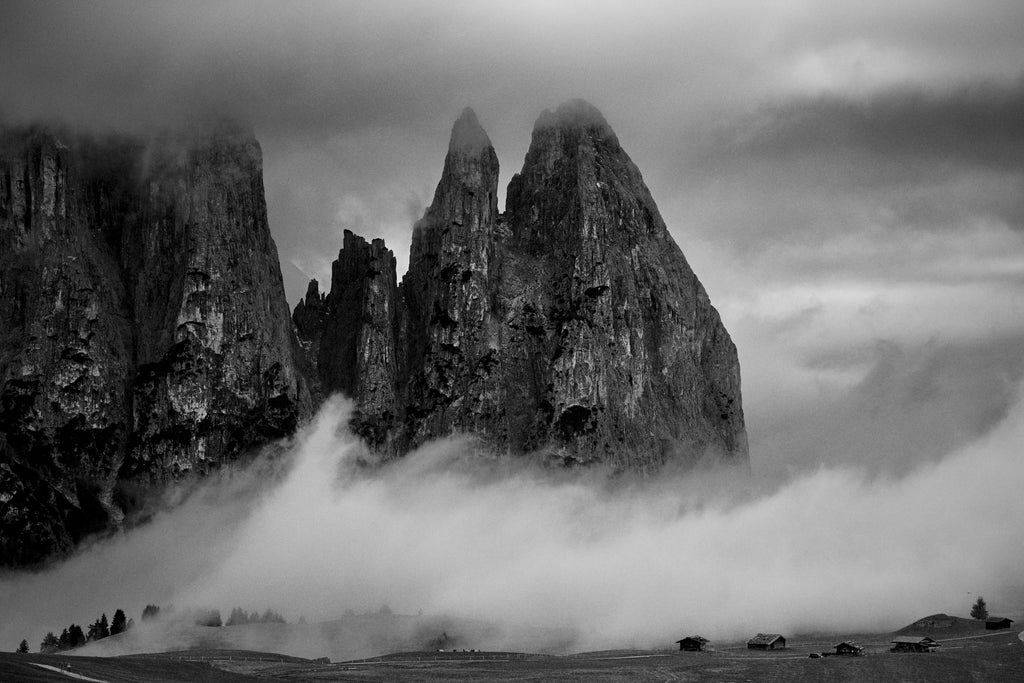 Photographic print of the Dolomites. A black and white mountain landscape of the jagged Sciliar massif peaks with mist swirling around the base and small alpine cabins in the foreground at Alpe di Siusi.