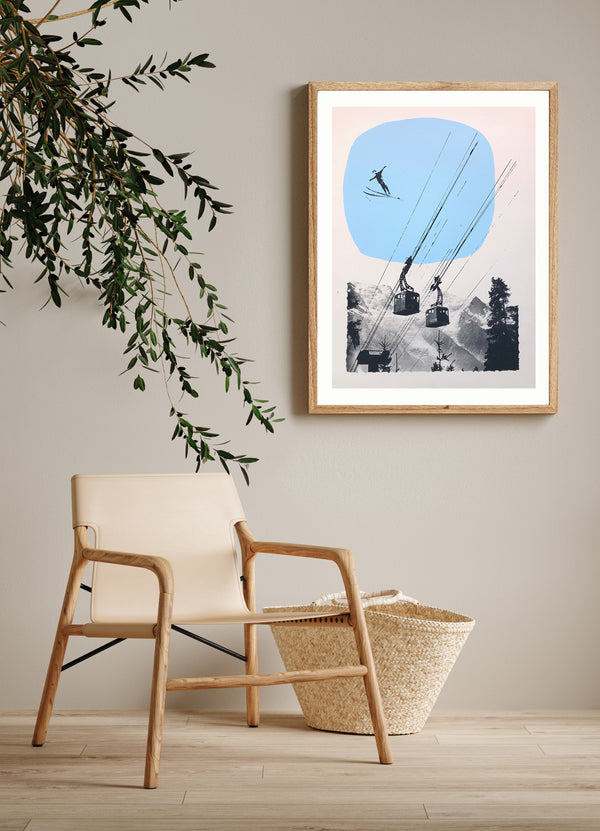 a framed retro sky poster style screen print showing a ski jumper and cable cars on a mountain backdrop with blue sky is on a wall beside a chair and a plant