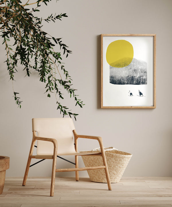 framed print with 2 cross country skiers against a mountain backdrop and huge yellow sun. In a room with a chair and plant